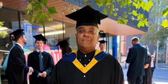 Image: Romesh stood in graduation cap and gown in front of LC building