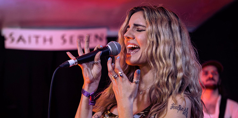 Mali Hâf with eyes closed singing into microphone on stage. Wearing her long, blonde, curly hair down and a Welsh themed vest top