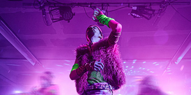 Students on stage wearing colourful attire with purple lighting. In School of Drama Production of A Midsummer Night's Dream